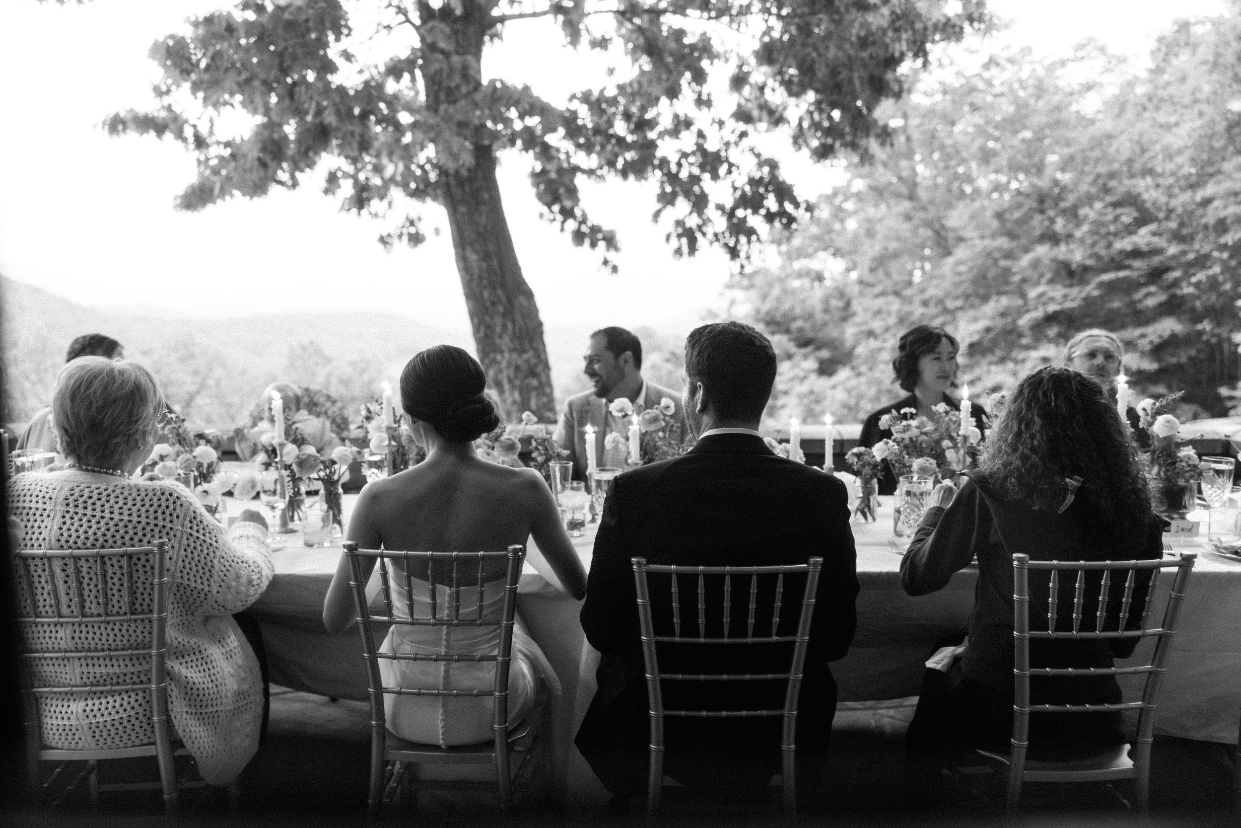 Wedding guests seated in white chairs enjoying an outdoor reception in black and white.
