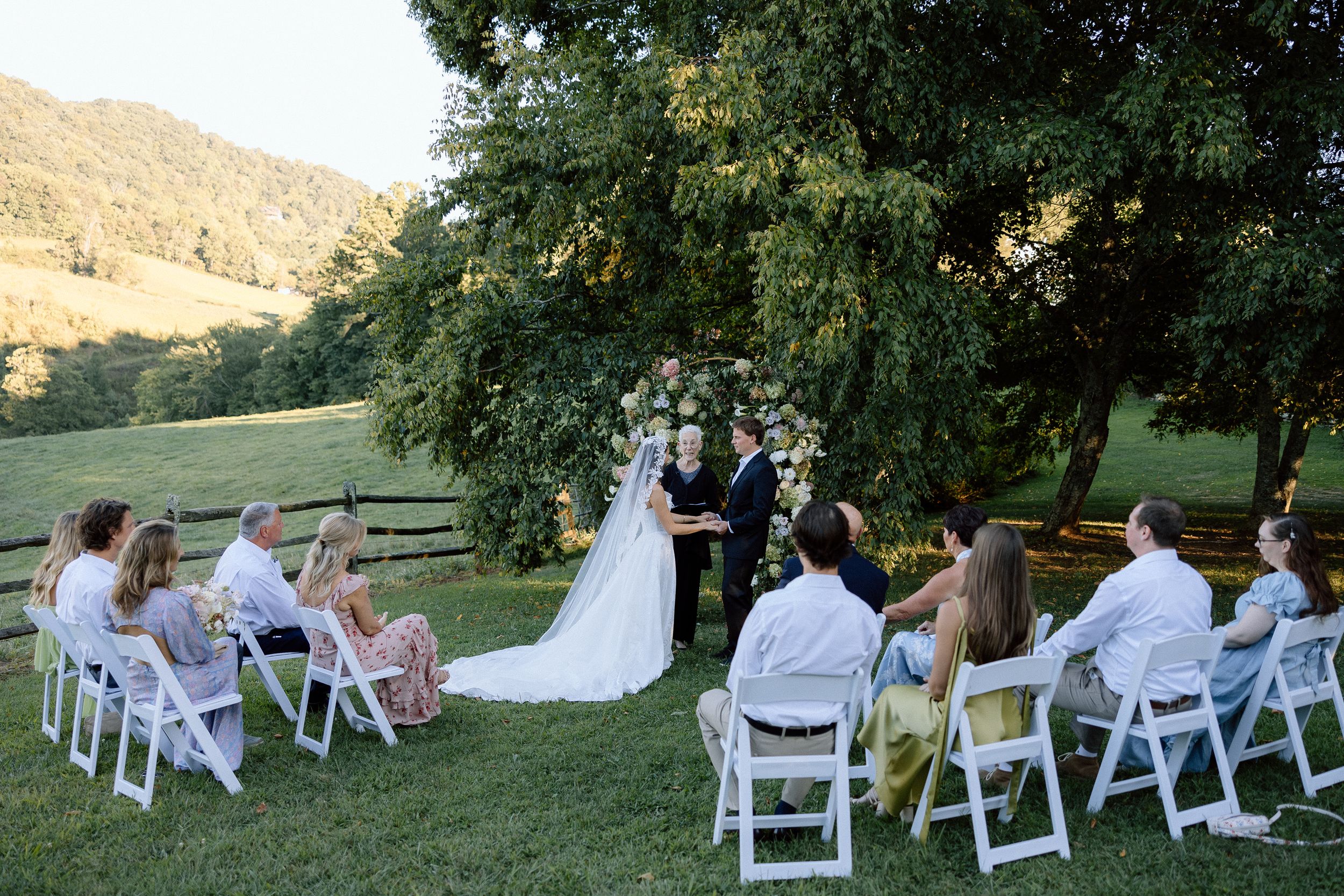 An intimate outdoor wedding ceremony takes place on a grassy hillside with white chairs arranged for guests.