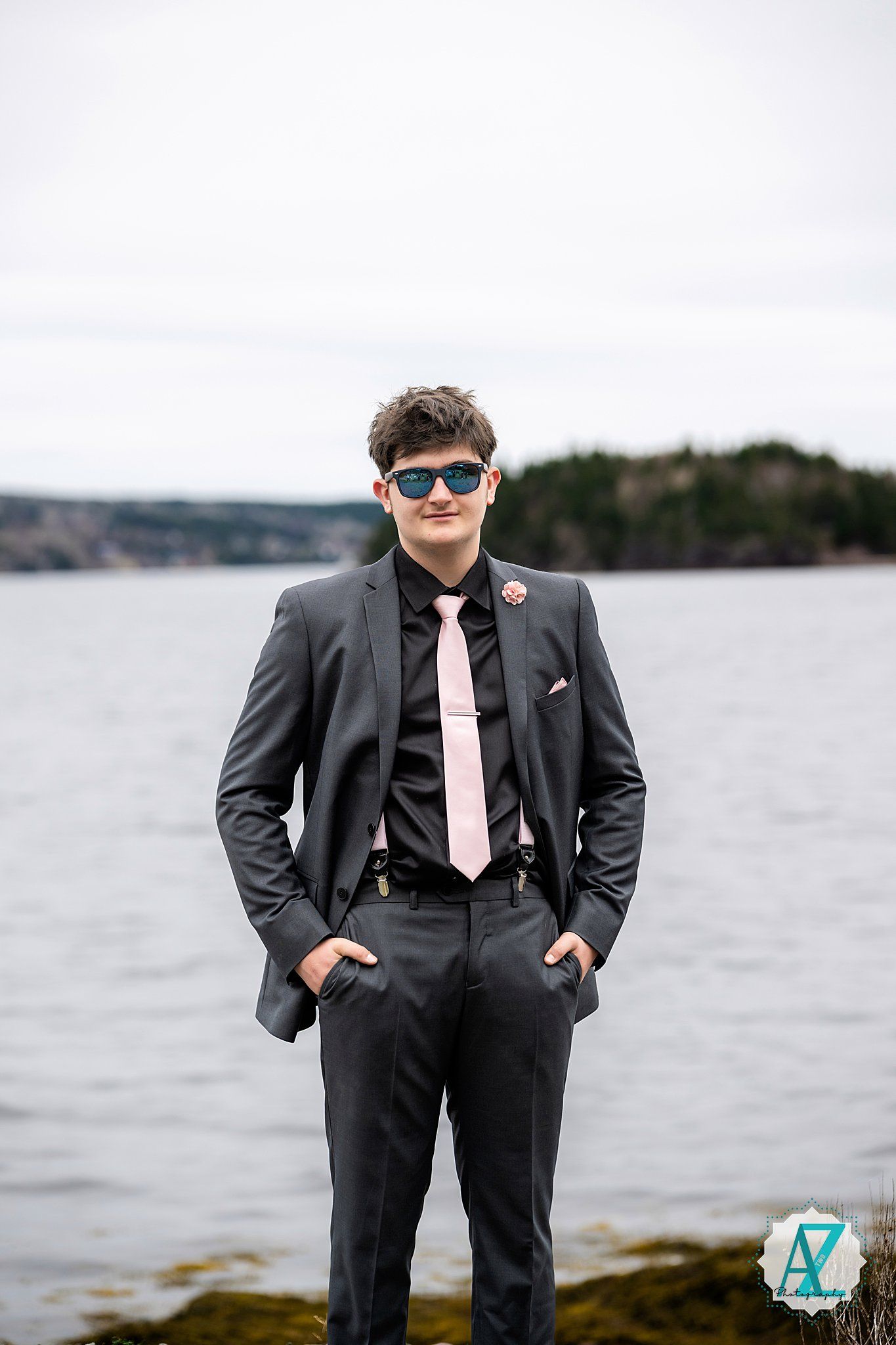 A person in a dark suit and pink tie stands confidently by a waterfront with sunglasses and hands in pockets.