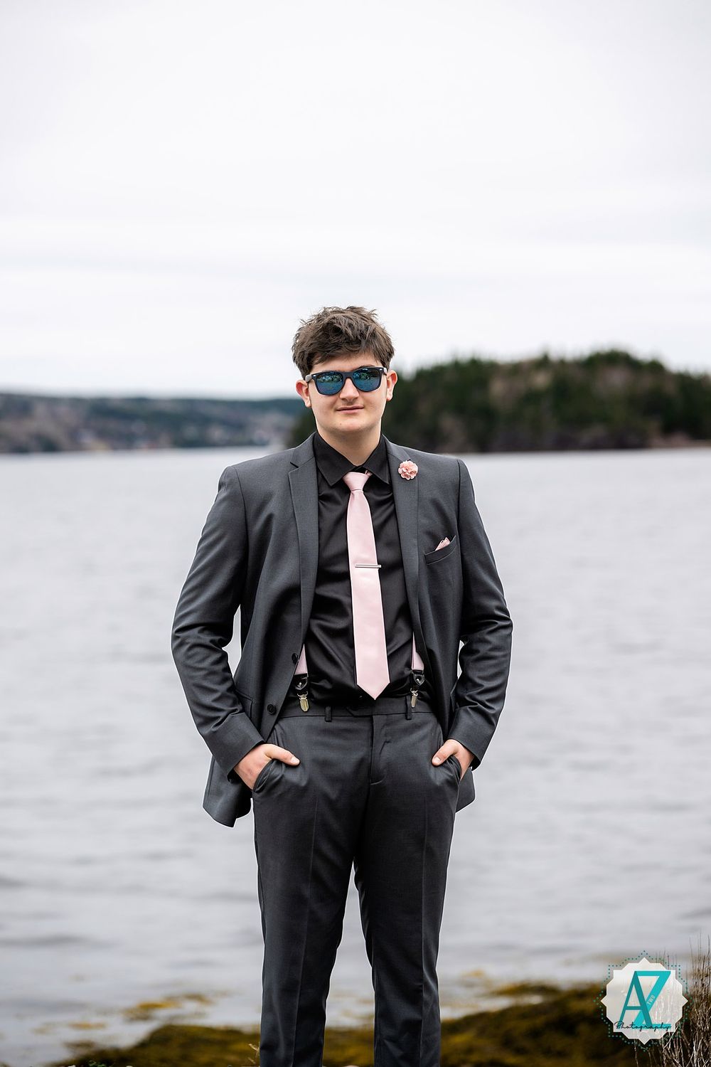 A person in a dark suit and pink tie stands confidently by a waterfront with sunglasses and hands in pockets.