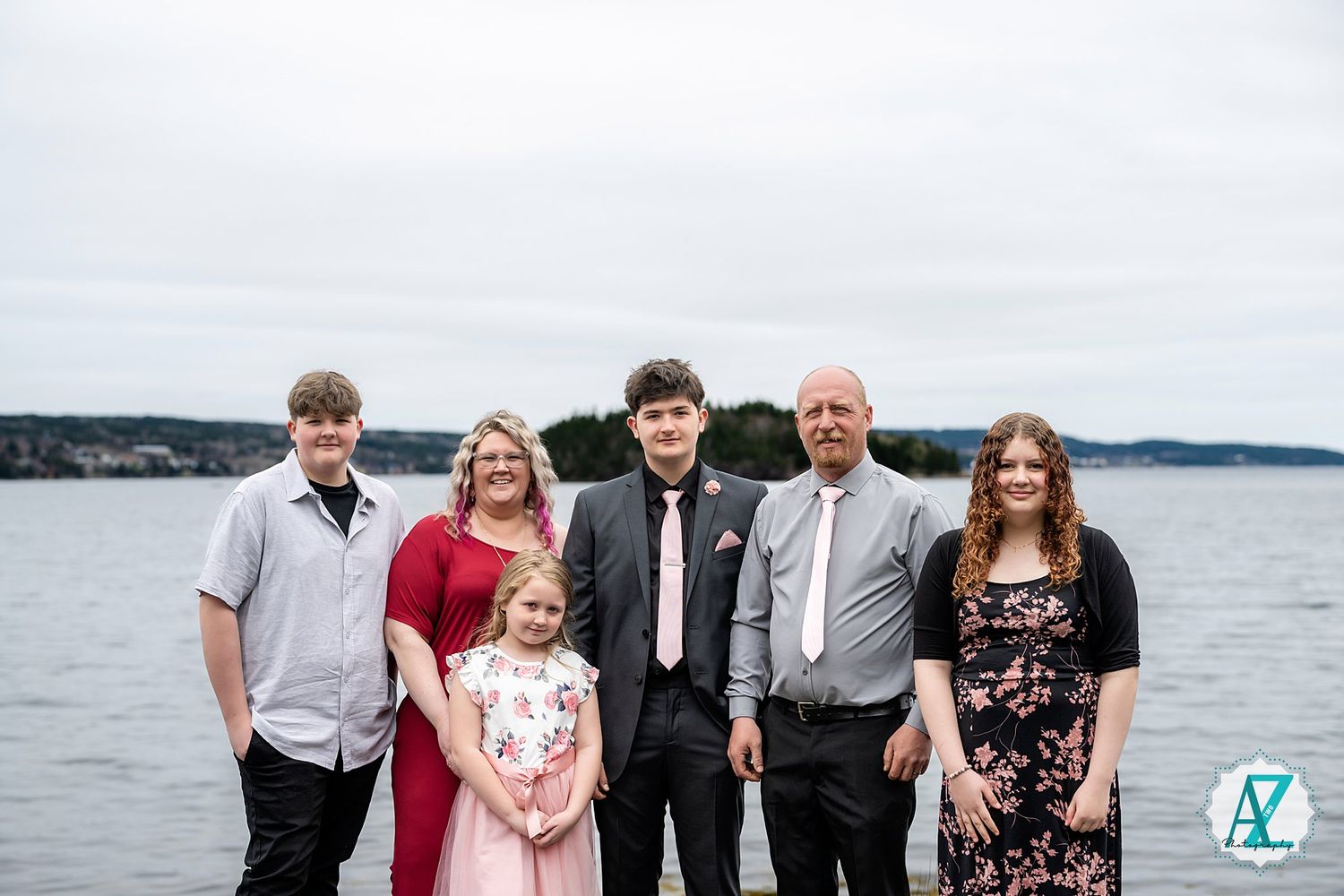 A group of formally dressed people stand together on a waterfront with a scenic lake and shoreline in the background.