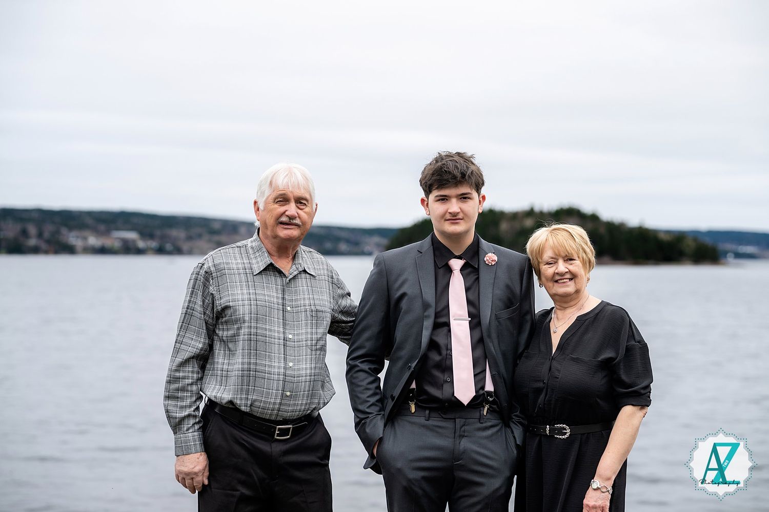 Three people stand together at a waterfront location with overcast skies and coastal scenery in the background.
