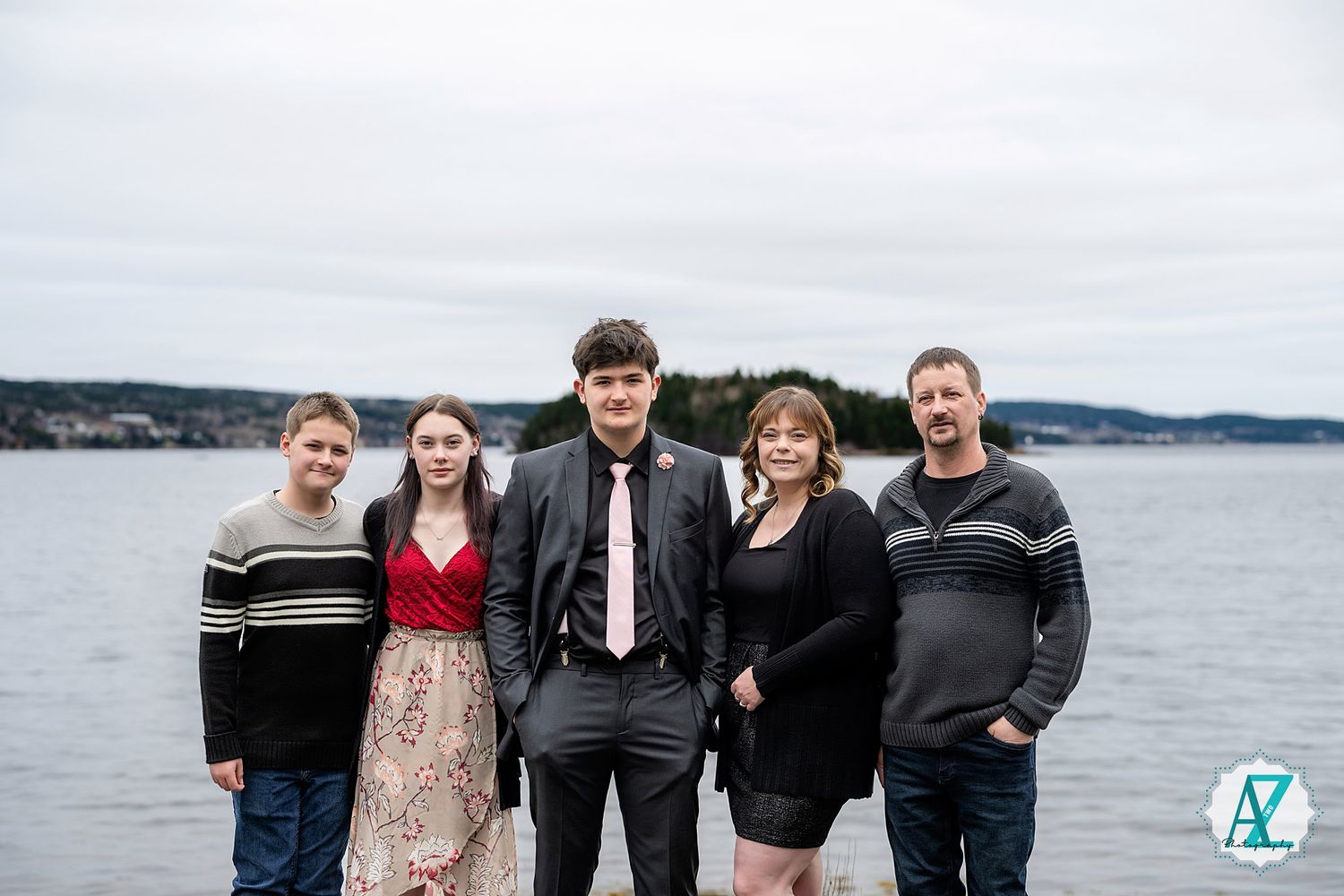 Five people in casual and formal attire stand together posing near a large body of water on an overcast day.
