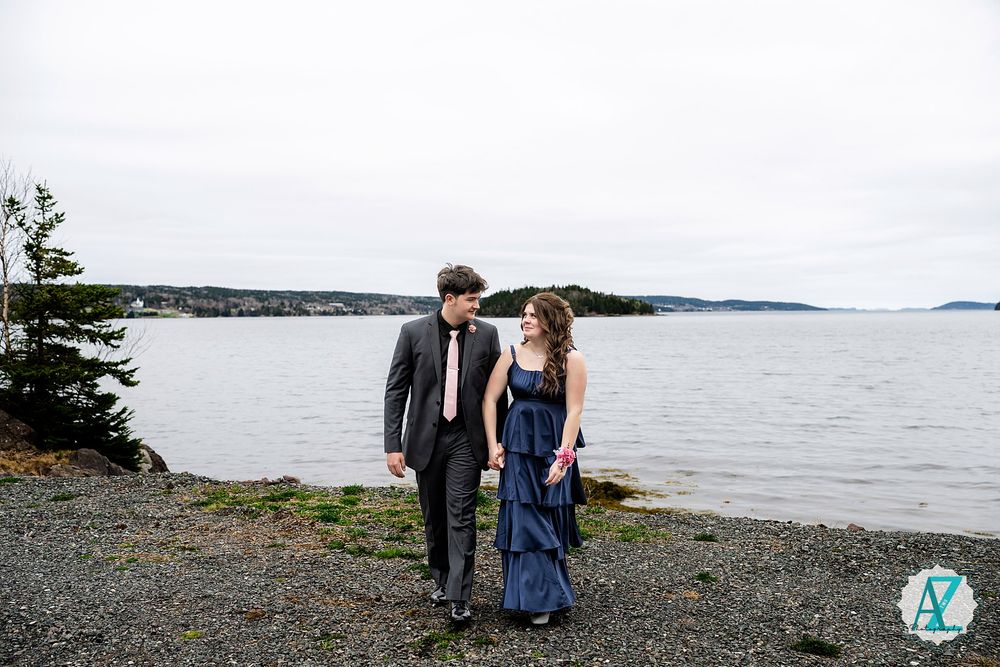A couple in formal attire walks along a rocky shoreline with a scenic waterfront and islands in the background.