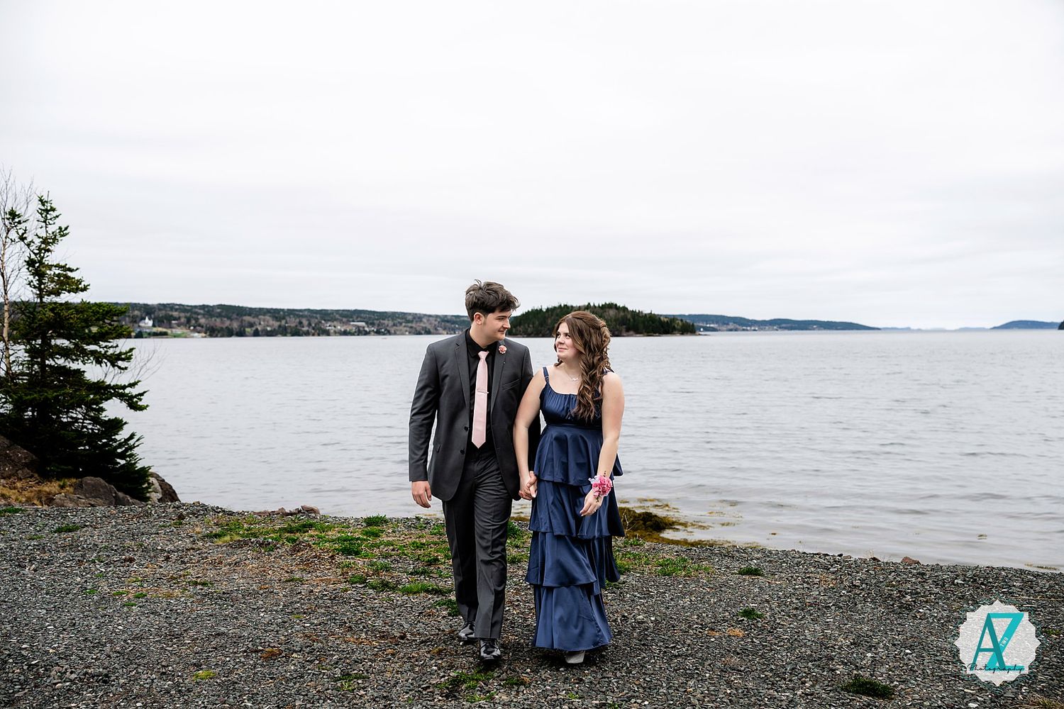 A couple in formal attire walks along a rocky shoreline with a scenic waterfront and islands in the background.