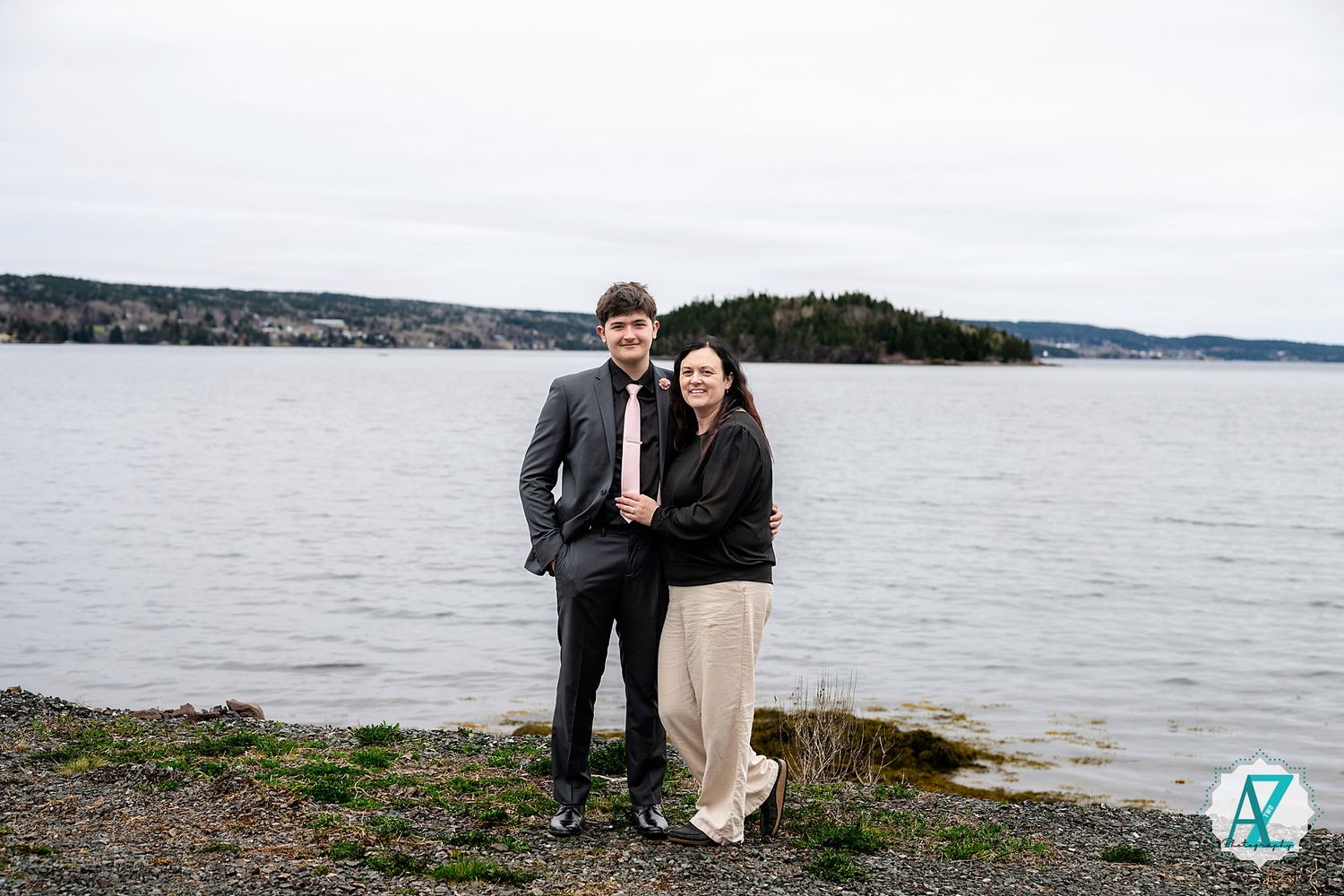 A couple stands together on a rocky shoreline overlooking a serene lake on an overcast day.