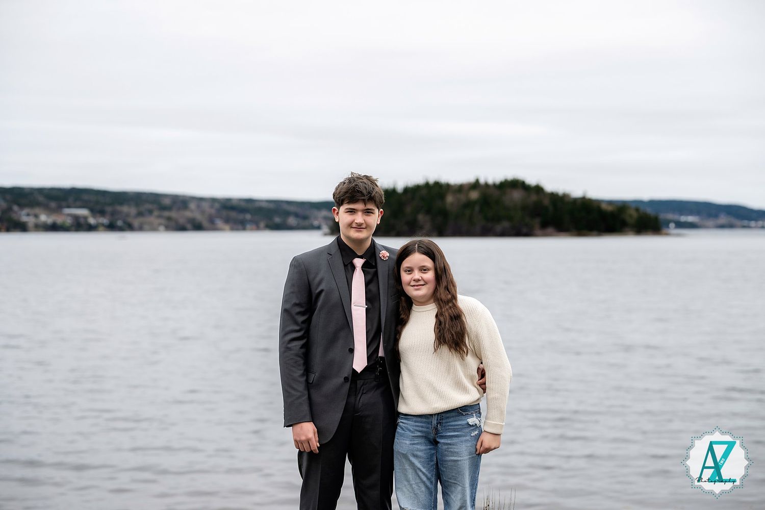 A couple stands together by a peaceful lake with hills in the background on an overcast day.