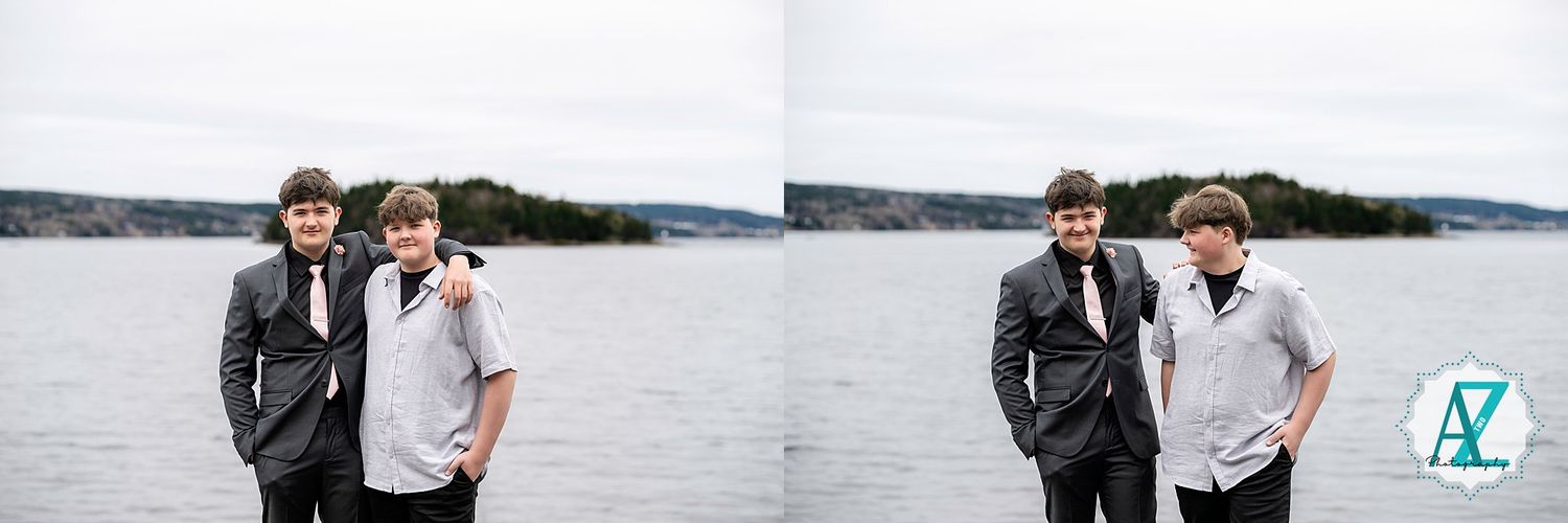 Two people in suits stand together by a lake on an overcast day against a wooded shoreline background.