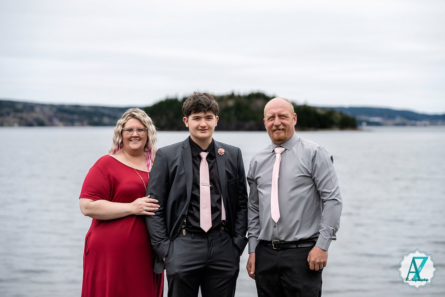 A family of three poses together near a scenic waterfront with rocky coastline and islands in the background.