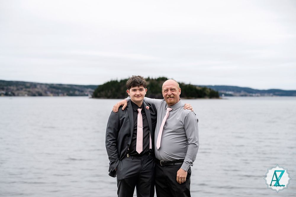 Two people stand together by a lake or ocean with hills in the background on an overcast day.