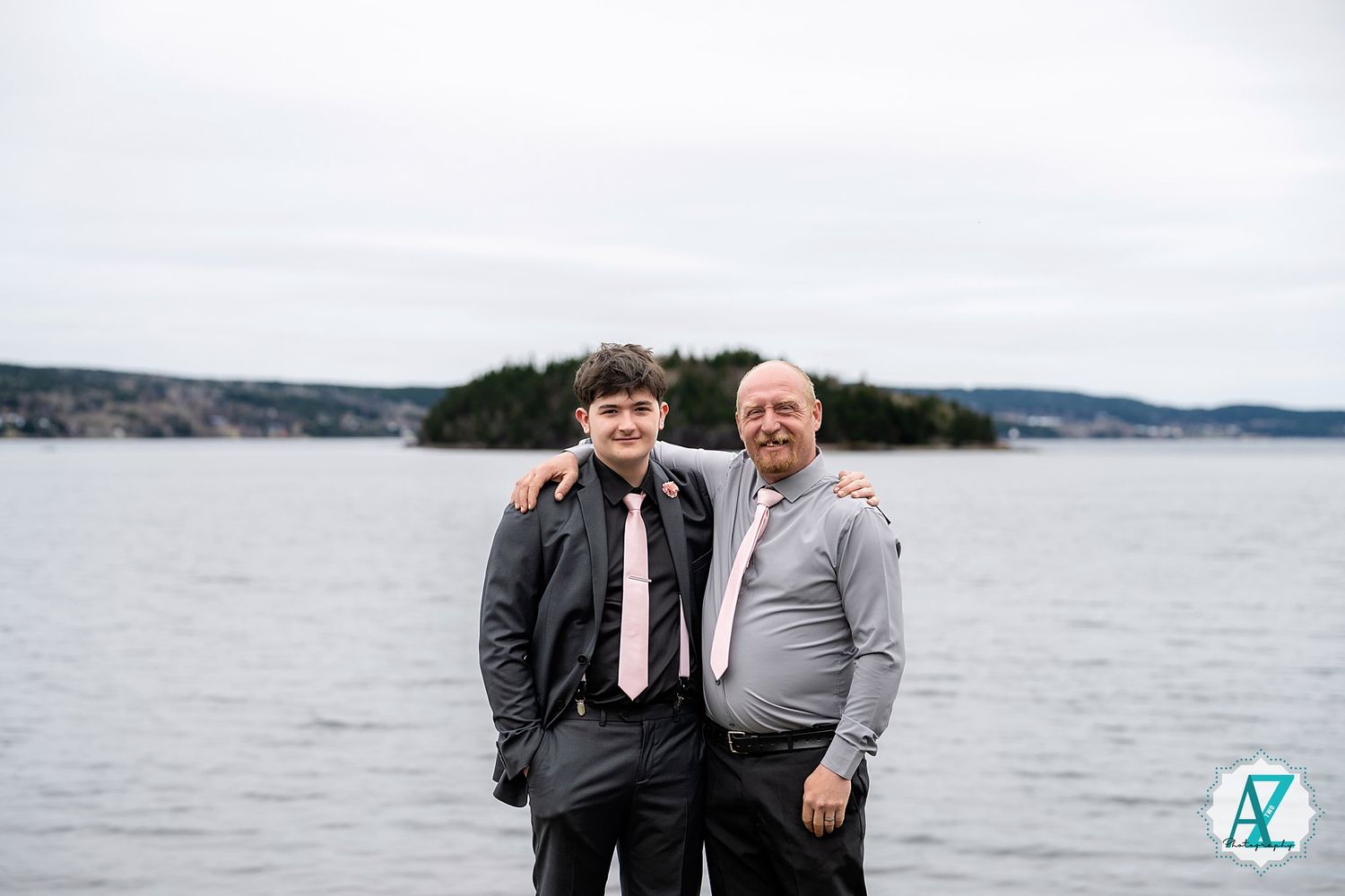 Two people stand together by a lake or ocean with hills in the background on an overcast day.