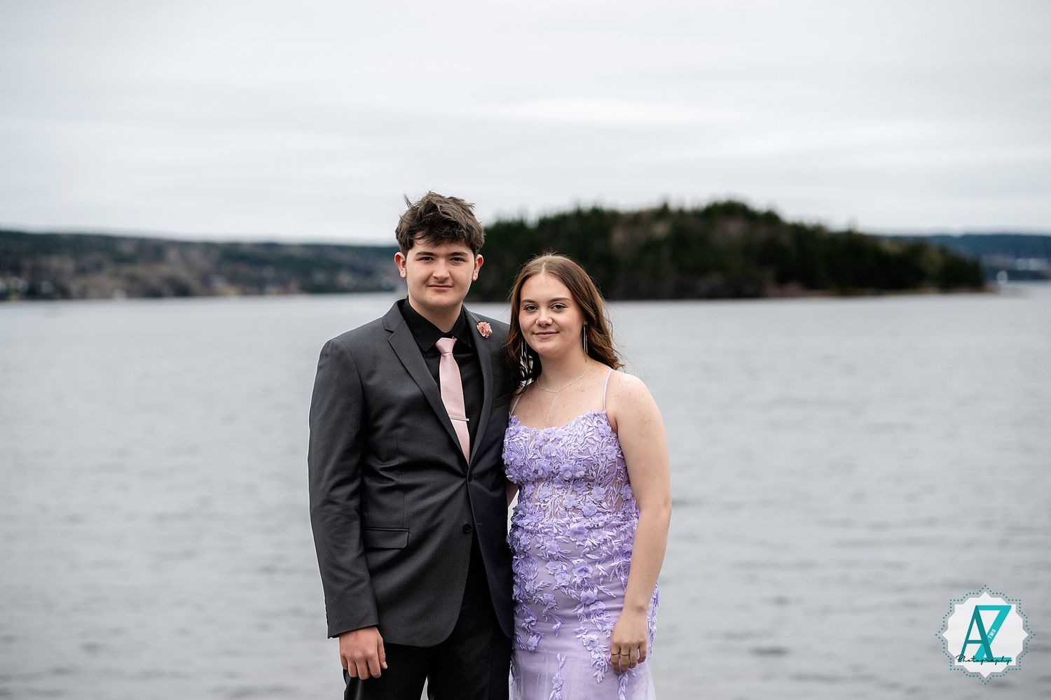 A couple in formal attire poses together with a water view and coastline in the background on an overcast day.