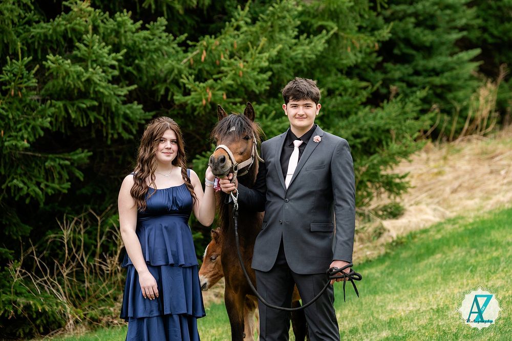 A couple stands outdoors with their horse amidst evergreen trees on a grassy hillside.