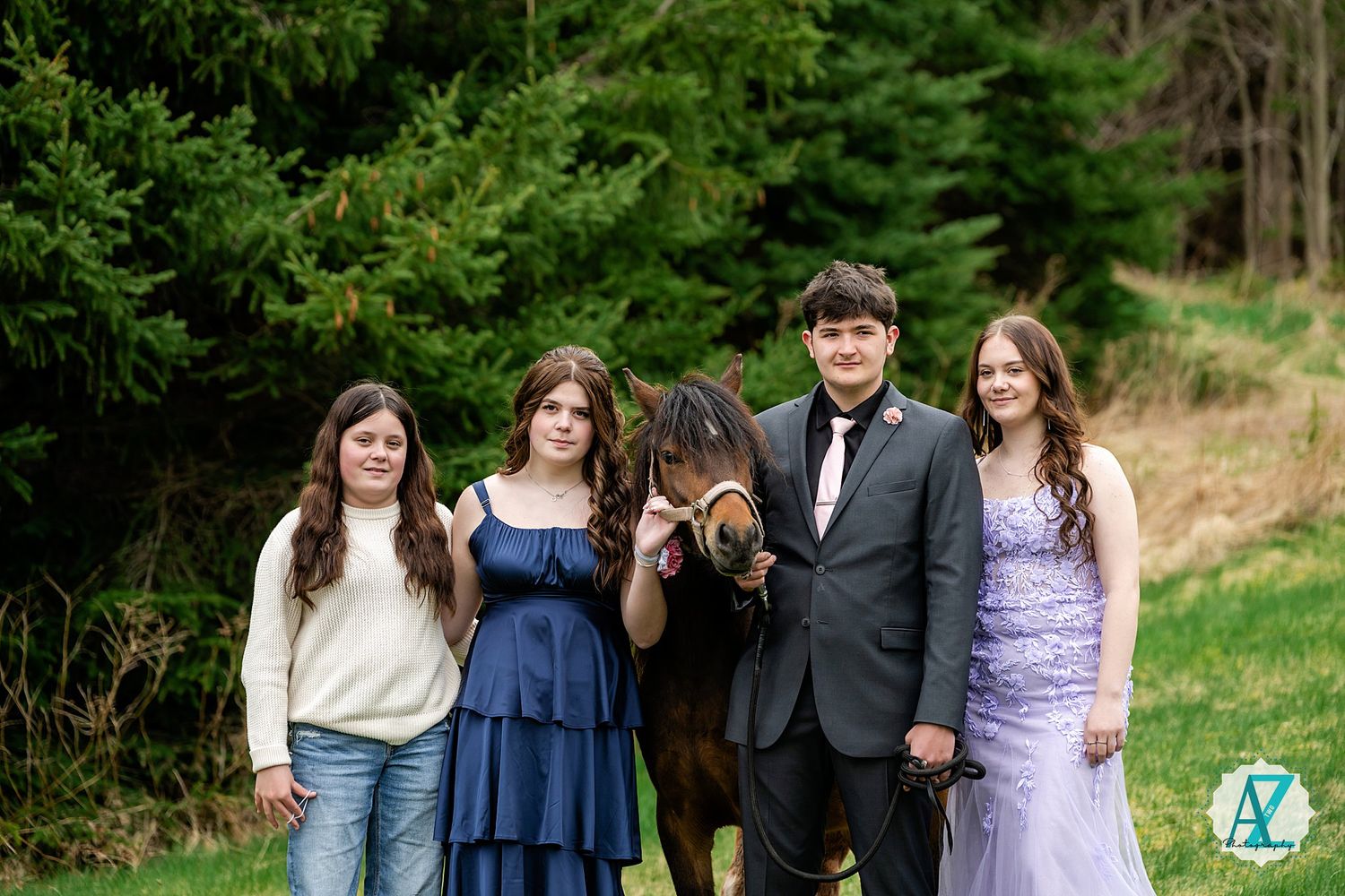 Teenagers in formal and casual attire pose together in front of evergreen trees in a grassy outdoor setting.
