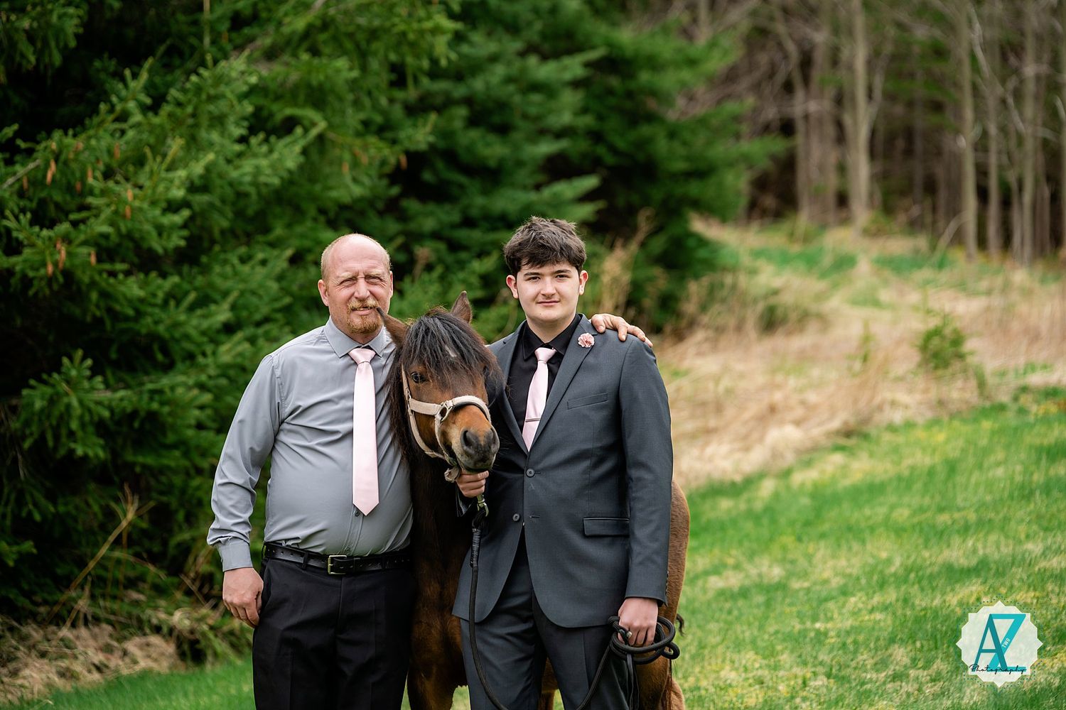 Two people pose with a brown horse on a grassy hill near evergreen trees and bare woods in the background.