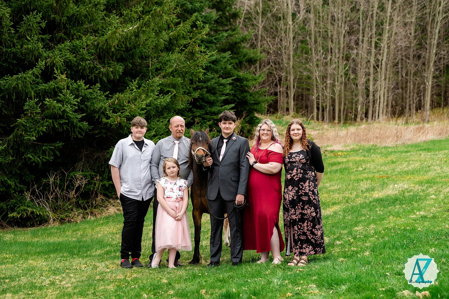 Small group dressed up for formal occasion standing together on grassy lawn with wooded forest backdrop.
