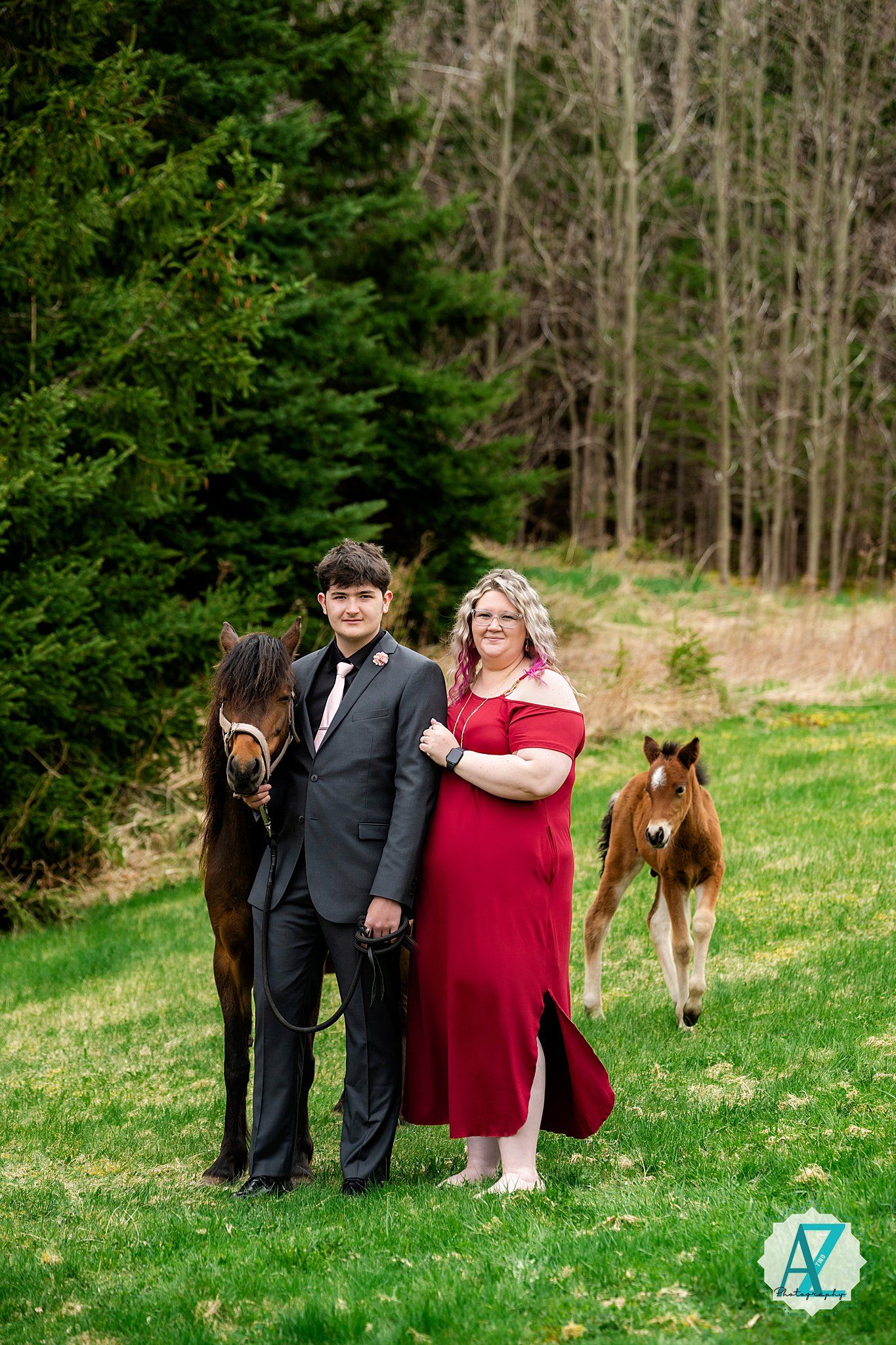 A couple in formal attire pose with horses on a grassy hillside with a small foal visible in the background.