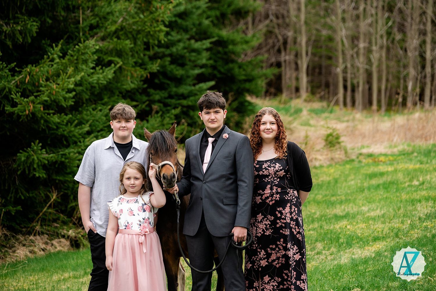 A family in formal attire poses together on a grassy hill with forest trees in the background.