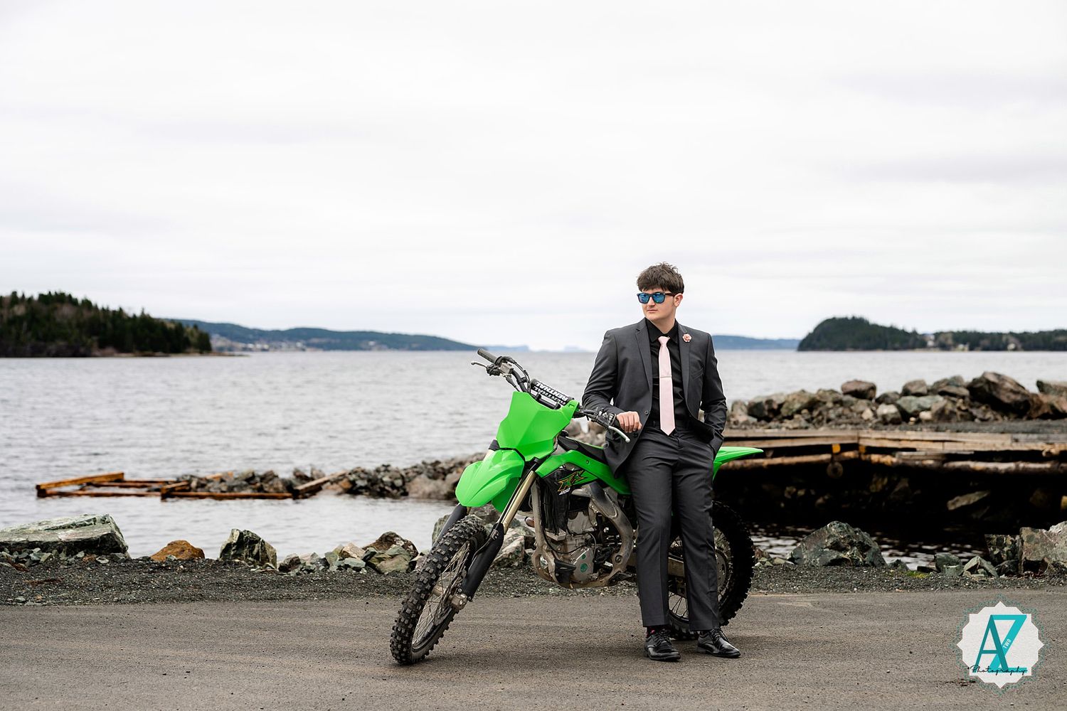 A bright green dirt bike and casually dressed figure stand on a rocky shore with water and islands in the background.