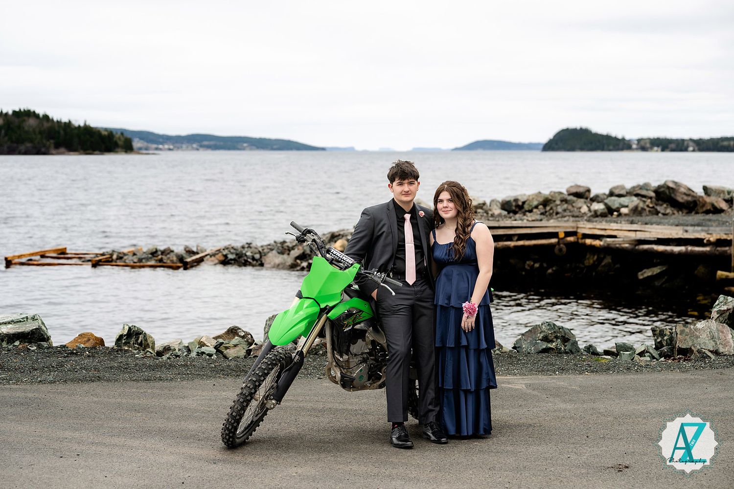 A couple poses with a bright green dirt bike against a coastal backdrop with rocks and water.