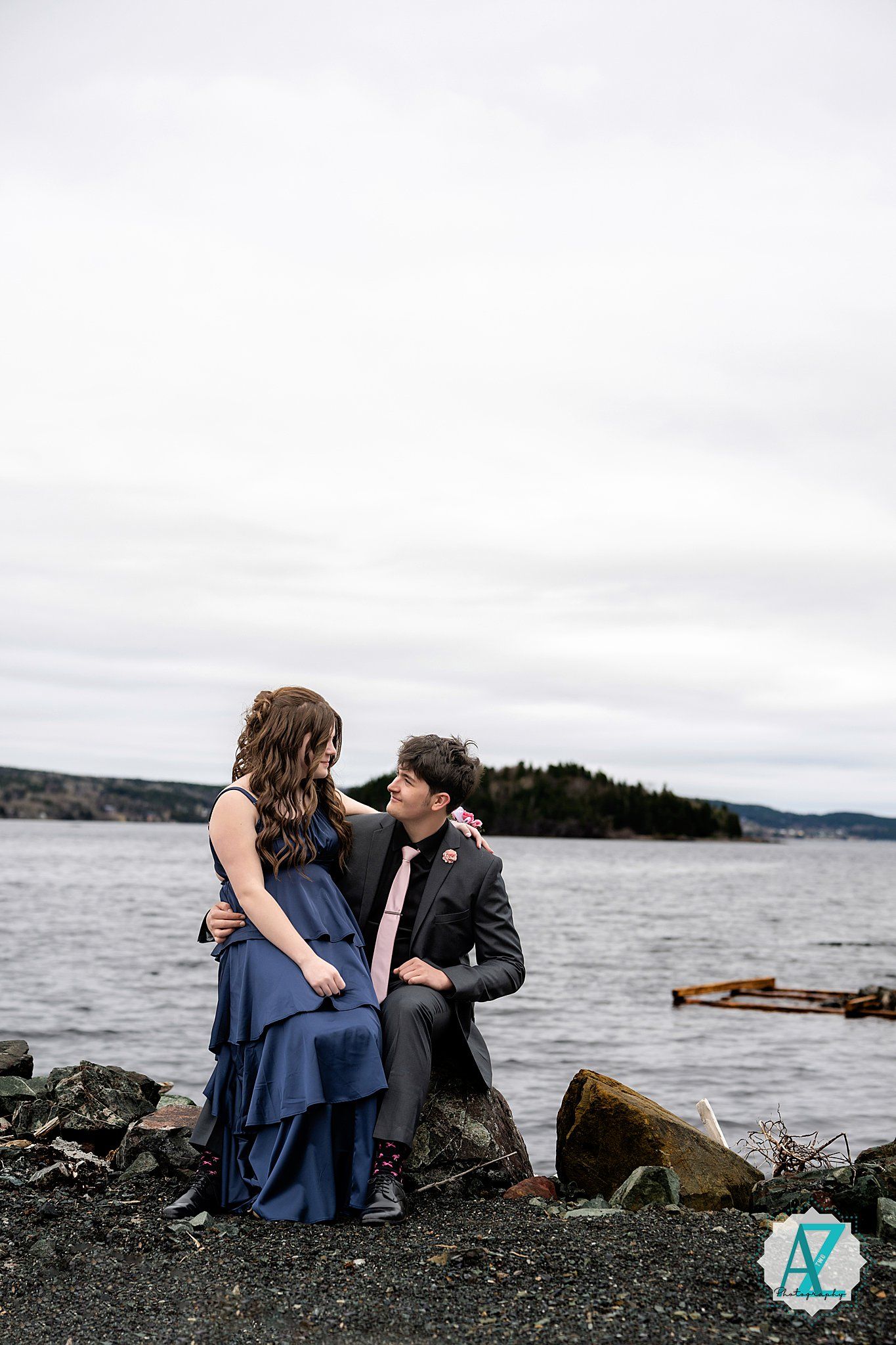 A couple in formal wear sits on rocky shoreline with water and overcast sky in the background during a photoshoot.