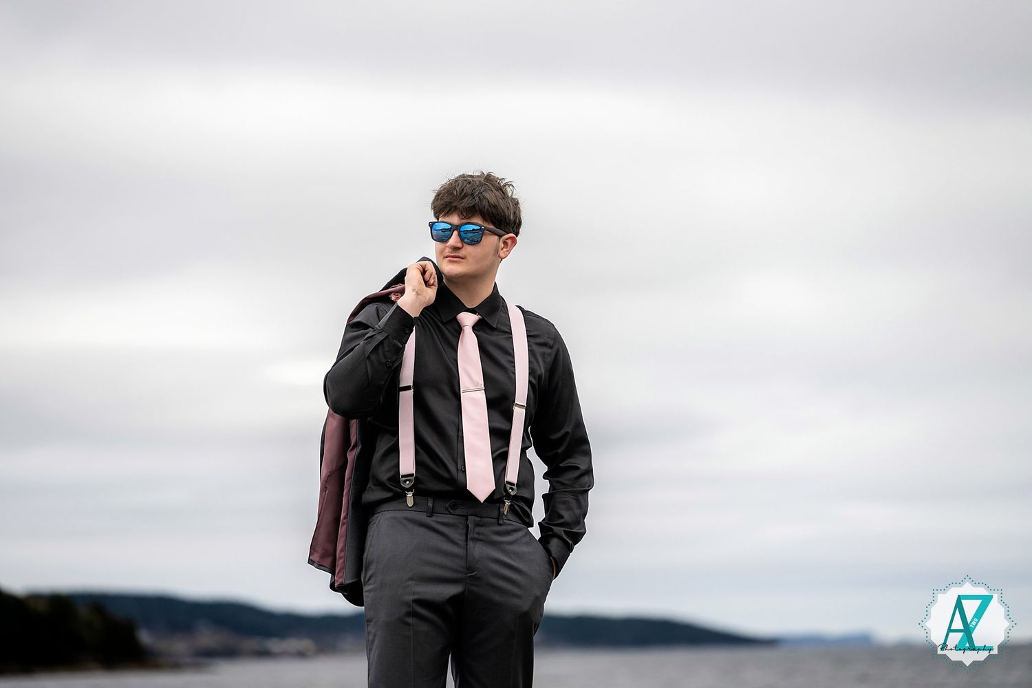 A person in formal attire with suit and tie stands on a hillside against an overcast sky.