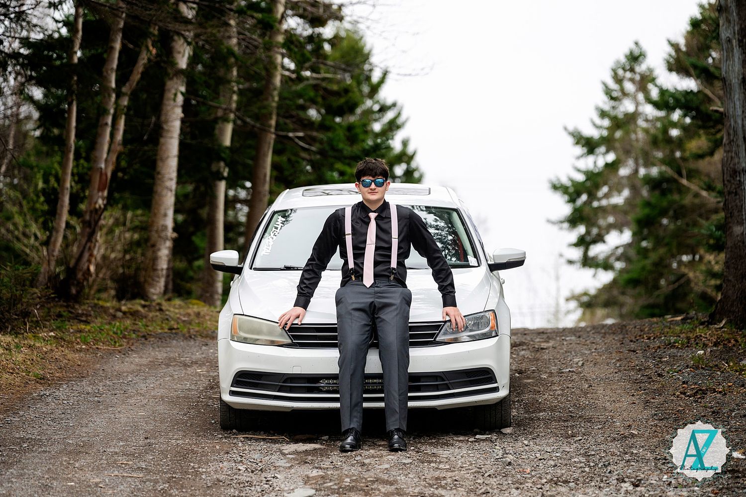 A white Volkswagen car parked on a dirt road surrounded by tall pine trees in a forest setting.