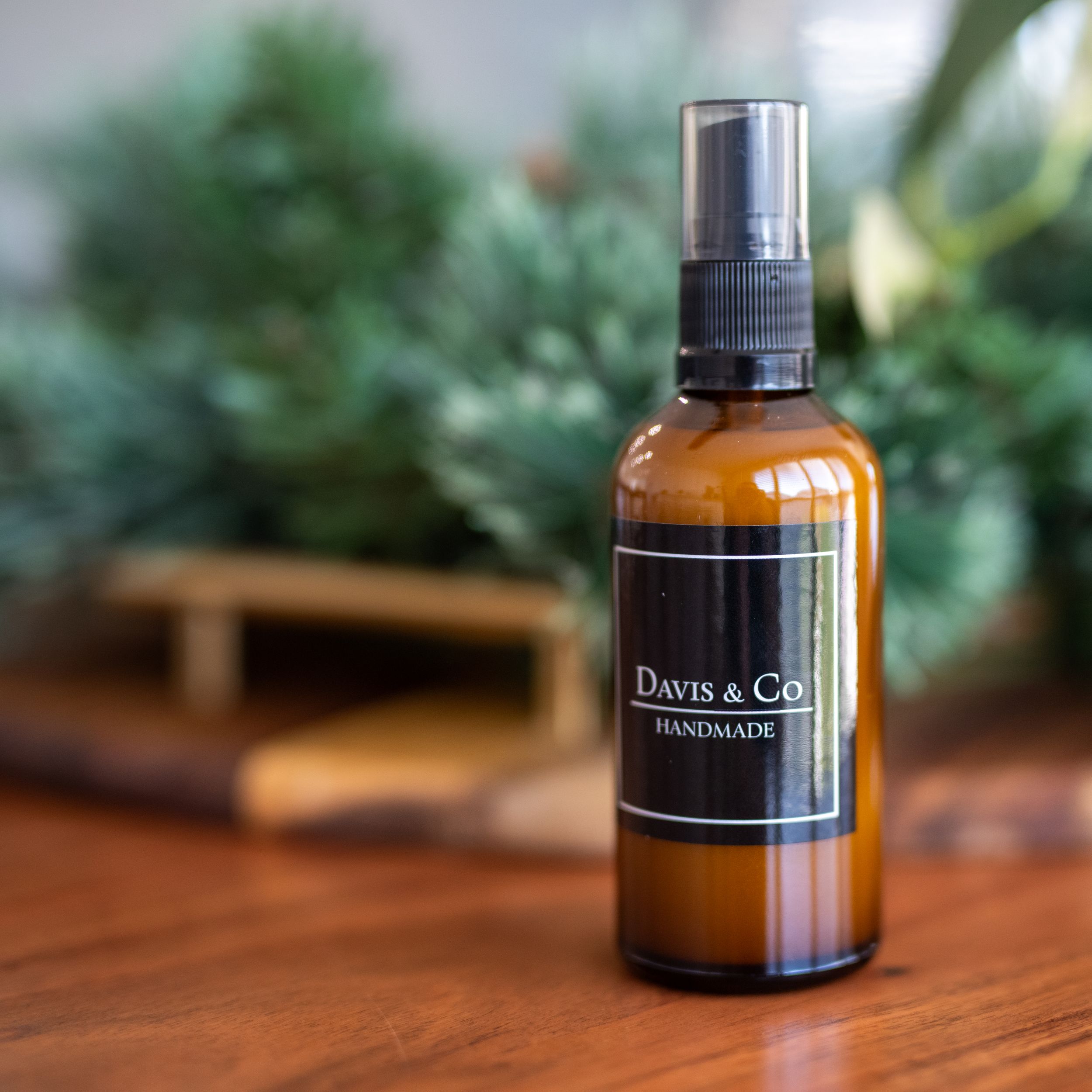 Amber glass bottle of Dawn & Co handmade product with spray nozzle sitting on wooden surface with green foliage background.
