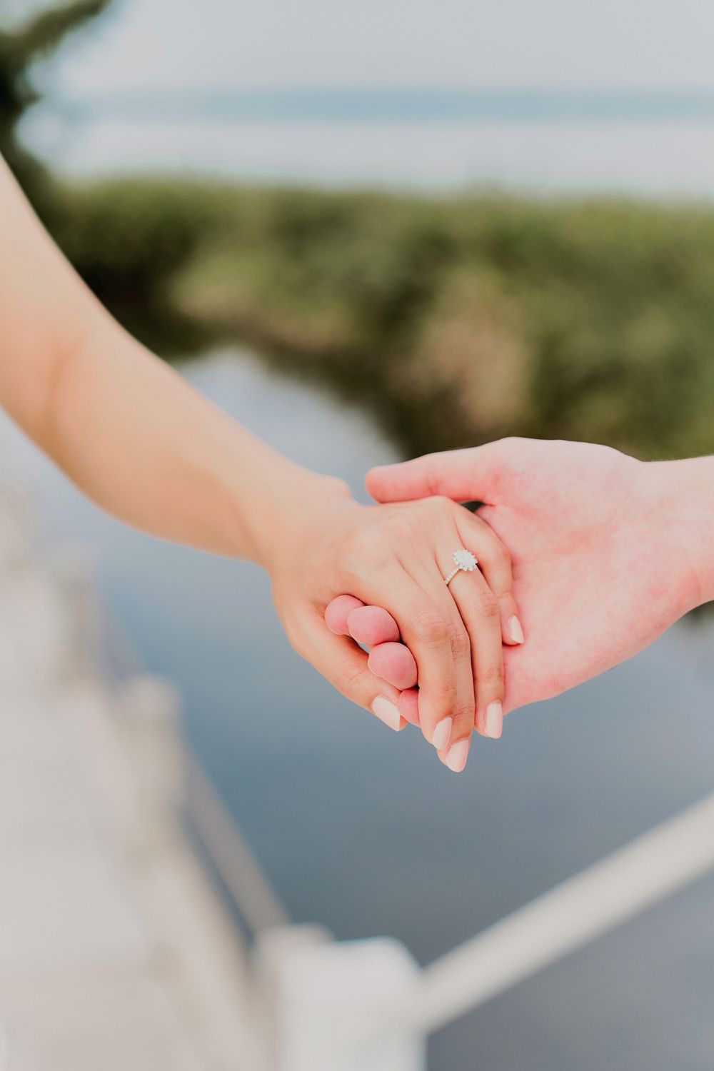 Close-up view of two hands holding each other with a wedding ring visible against a blurred outdoor background.