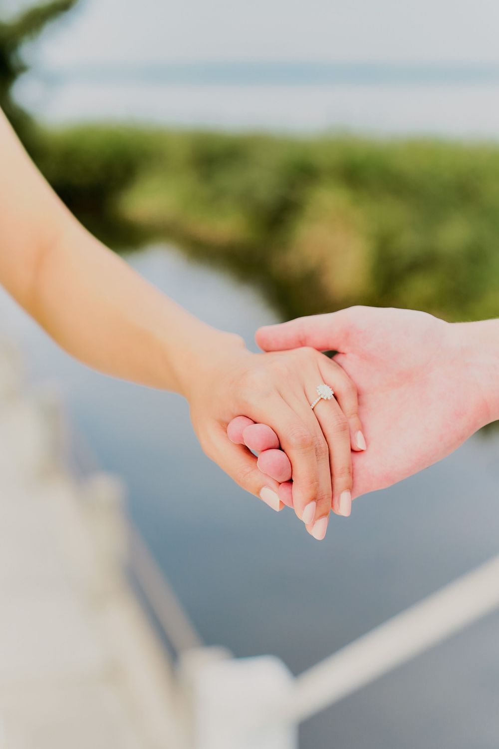 Close-up view of two hands holding each other with a wedding ring visible against a blurred outdoor background.