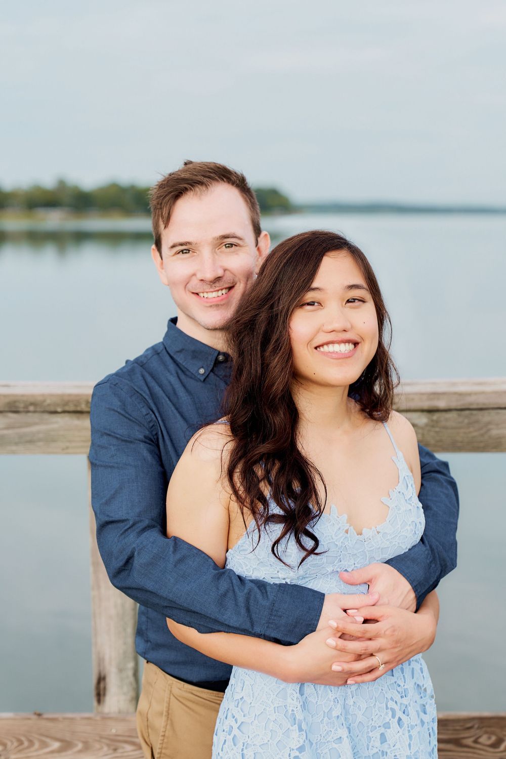 A loving couple embraces on a wooden dock overlooking a serene lake on a peaceful overcast day.