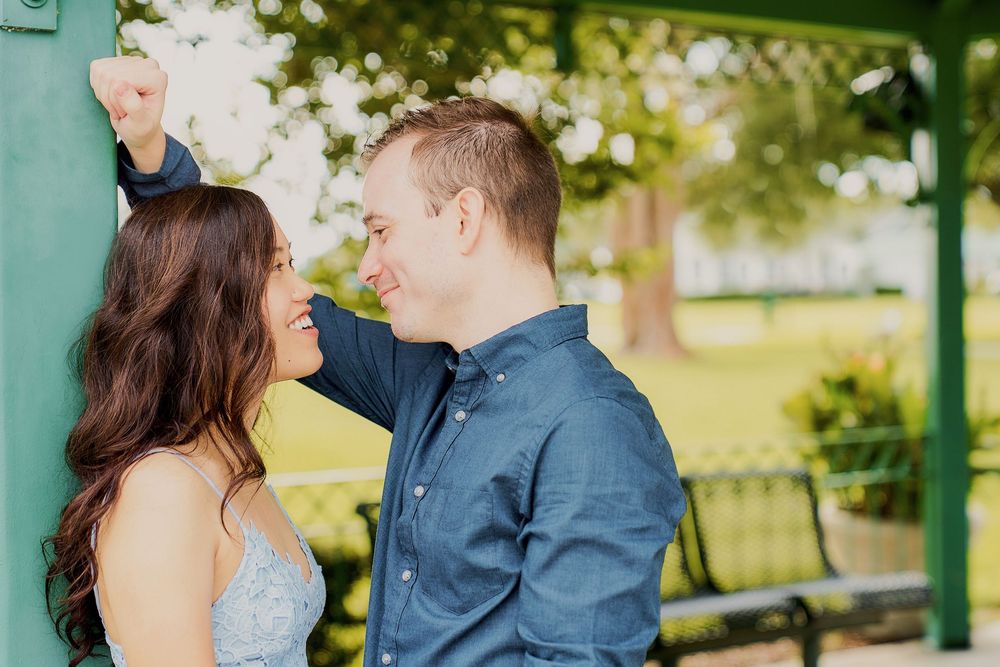 A romantic couple shares a tender moment in a park gazebo with blooming trees in the background on a sunny day.