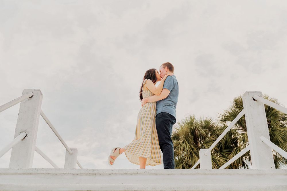 A couple embraces romantically on a white bridge with palm trees and soft clouds in the background.