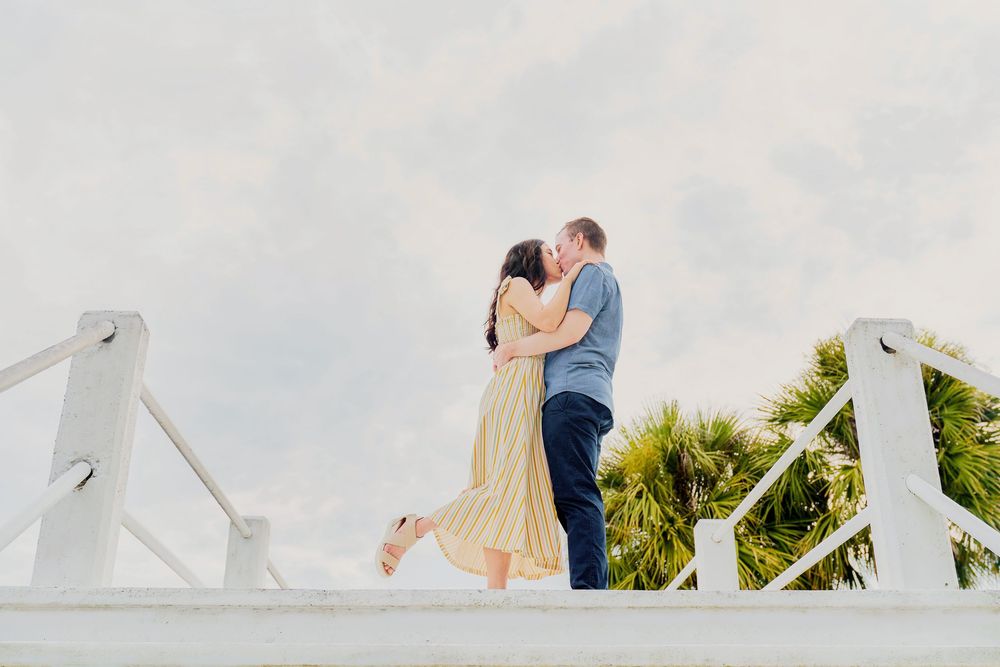 A couple embraces romantically on a white bridge with palm trees and soft clouds in the background.