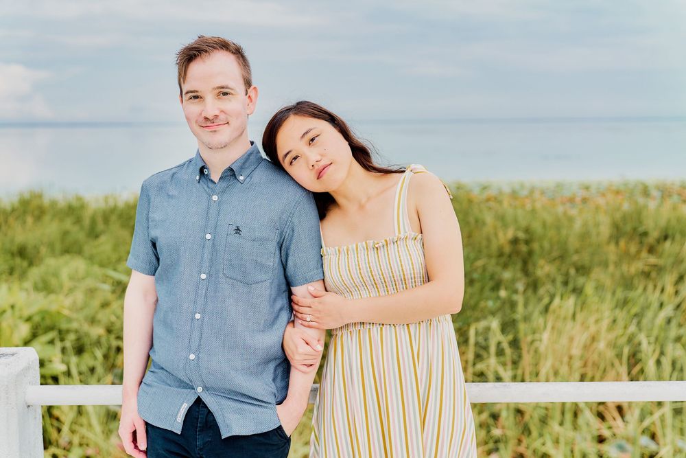 A couple poses together near a white fence on a sunny day with green grass and foliage in the background.