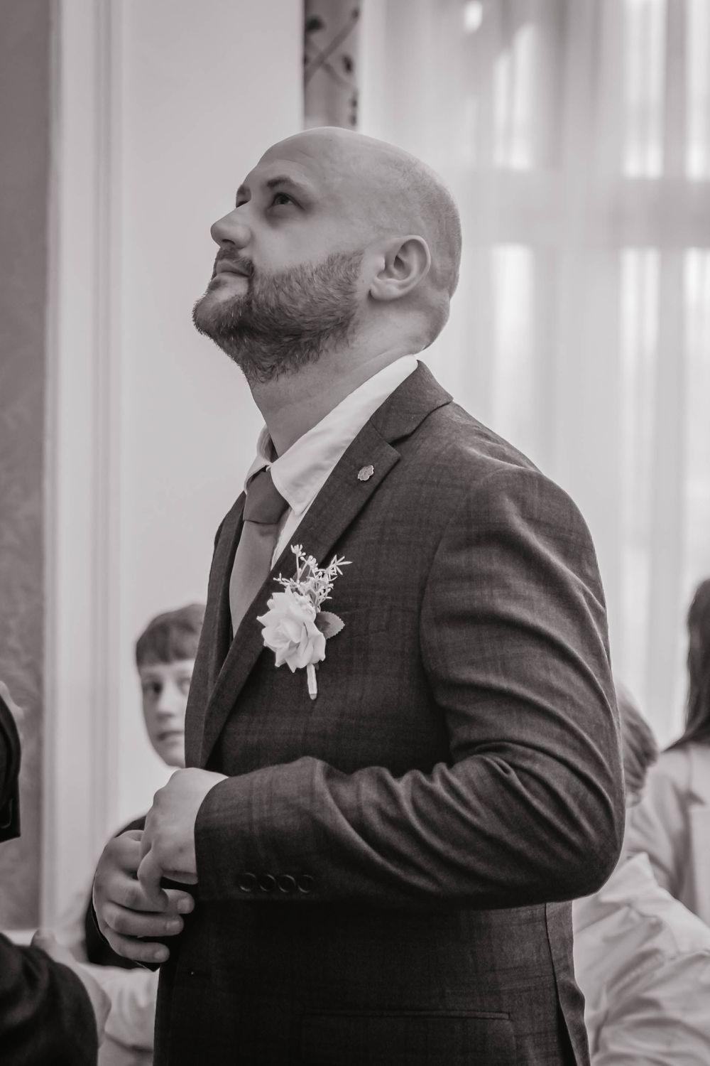 Black and white photo of groom in suit with boutonniere looking up during emotional wedding ceremony moment.