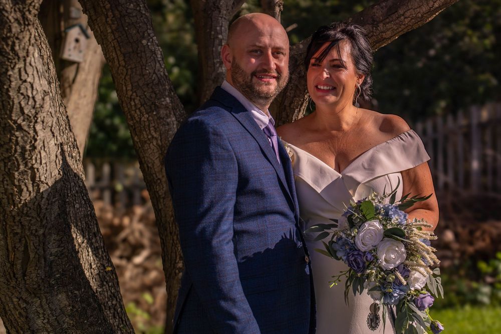 A couple on their wedding day poses under a tree with the bride holding a purple and blue floral bouquet.