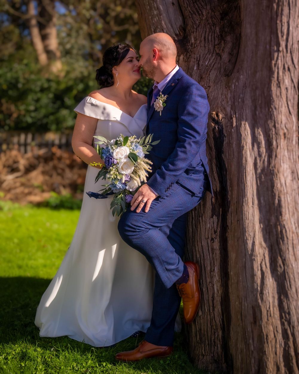A couple shares a romantic moment against a tree trunk at their outdoor wedding, with the bride holding a purple floral bouquet.