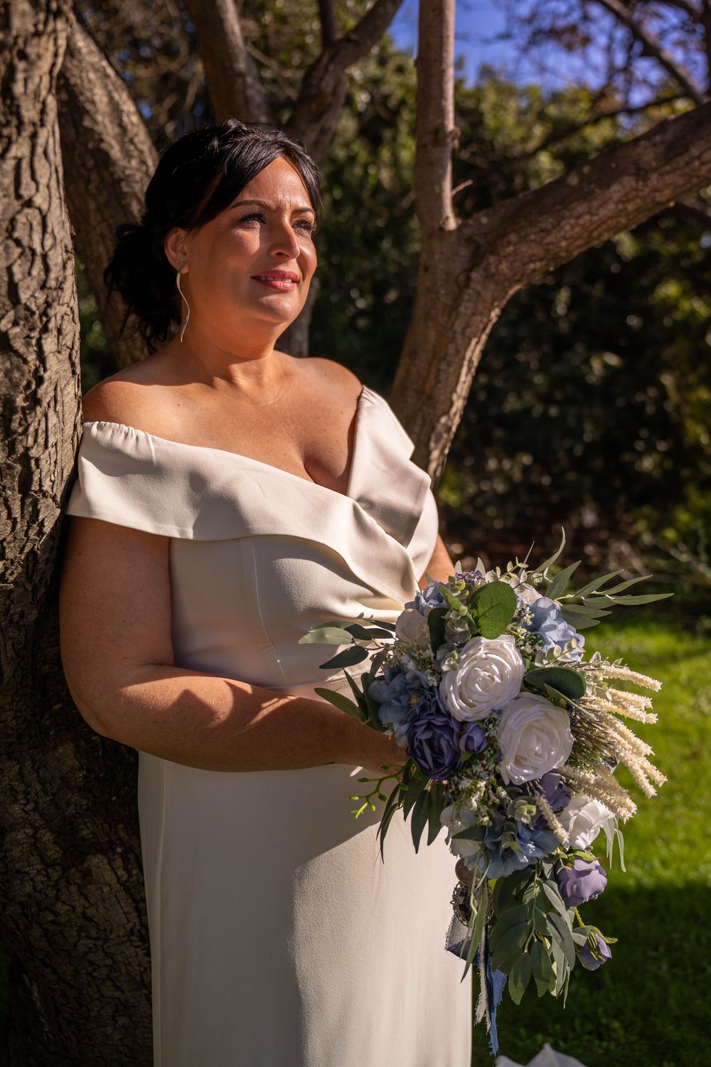 A bride in an elegant off-shoulder white gown holds a blue and white floral bouquet while standing by a tree in sunlight.