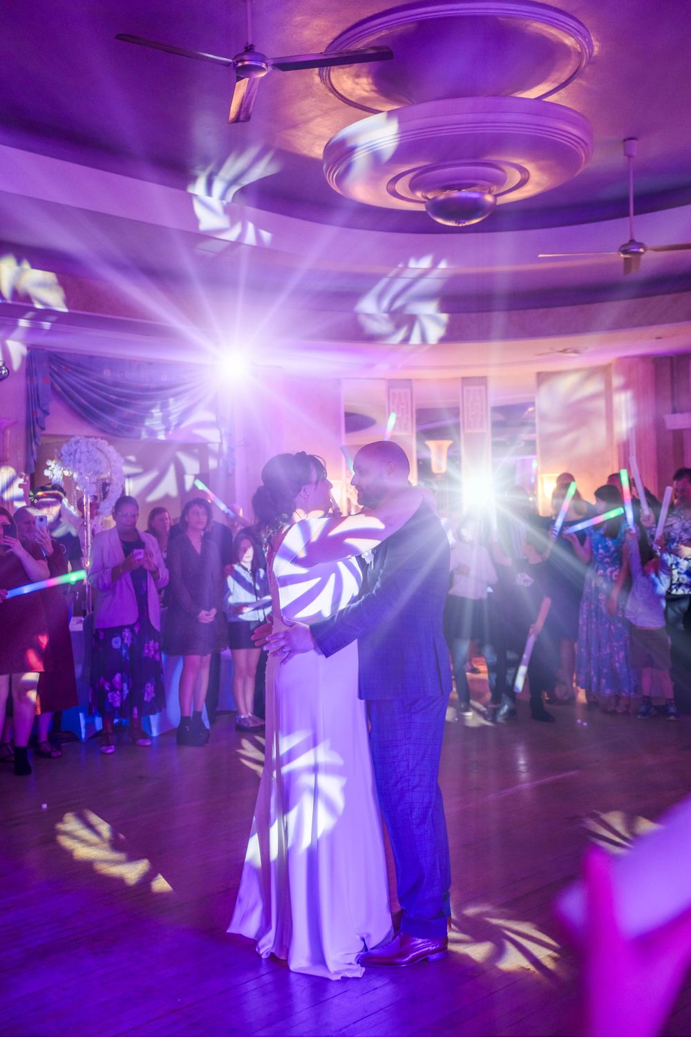 A couple dances in a purple-lit ballroom with swirling light patterns during what appears to be a wedding reception.