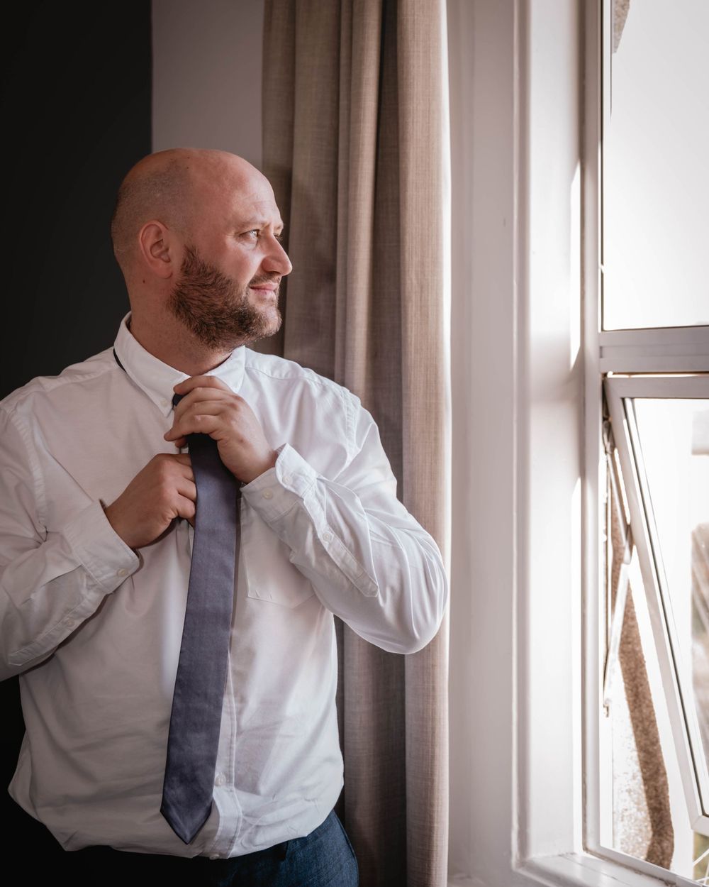 Professional in white dress shirt and gray tie adjusts collar while gazing thoughtfully out window in natural light.