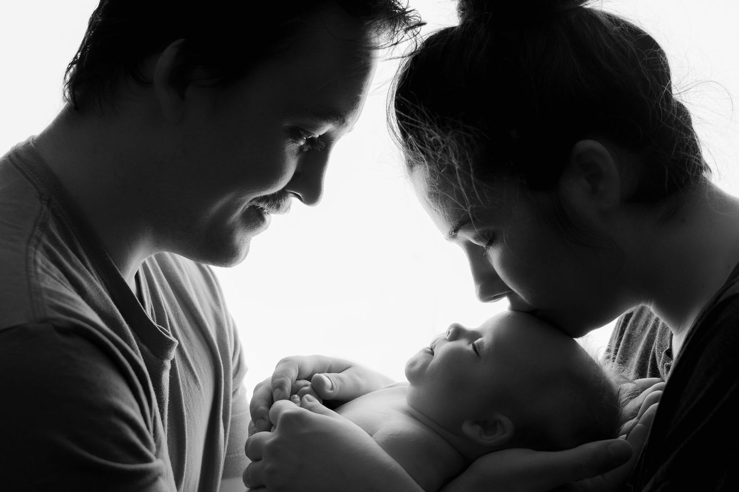 Black and white photo of two parents lovingly gazing down at their newborn baby in a tender family moment.
