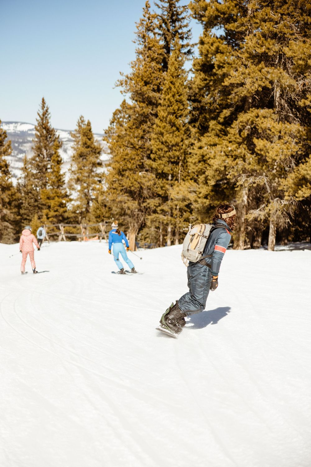 Snowboarder rides down slope with pine trees lining snowy mountain run.