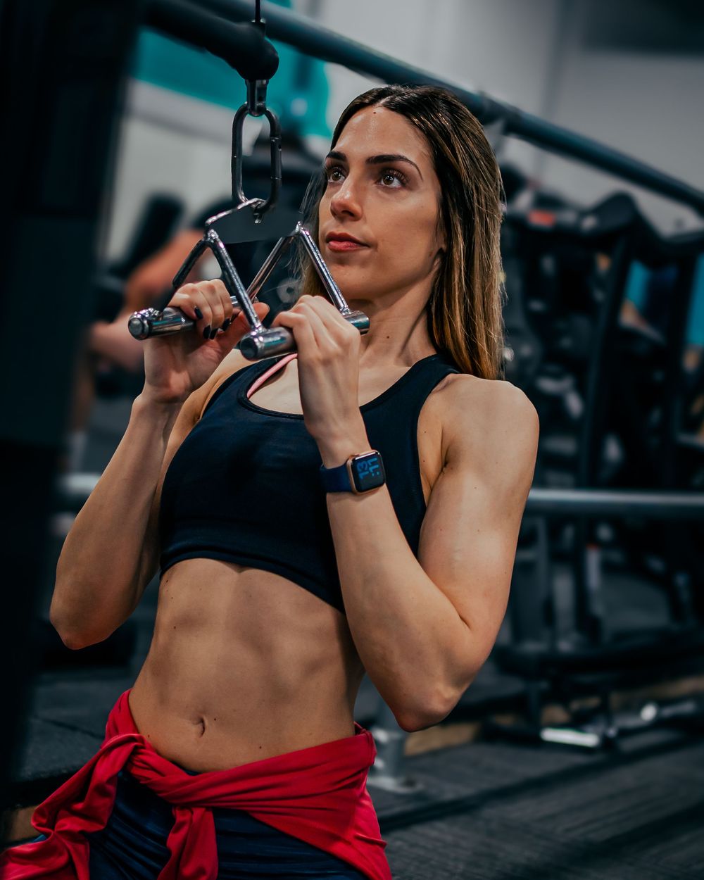 Athletic woman performing lat pulldown exercise at gym in black sports bra and red tied shirt.