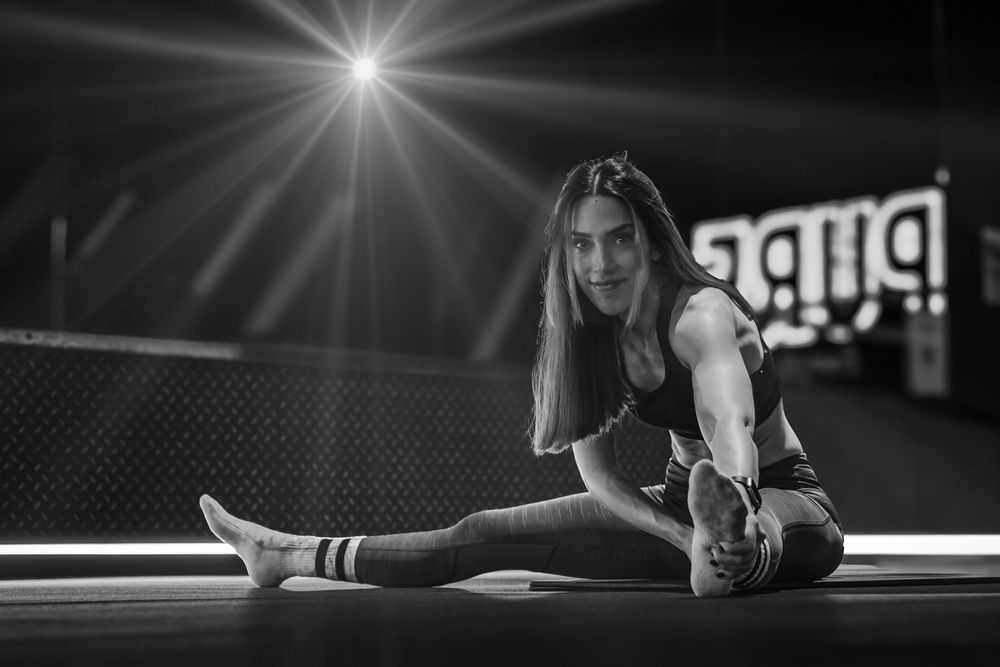 Black and white capture of stretching exercise with bright overhead lighting in gym setting.