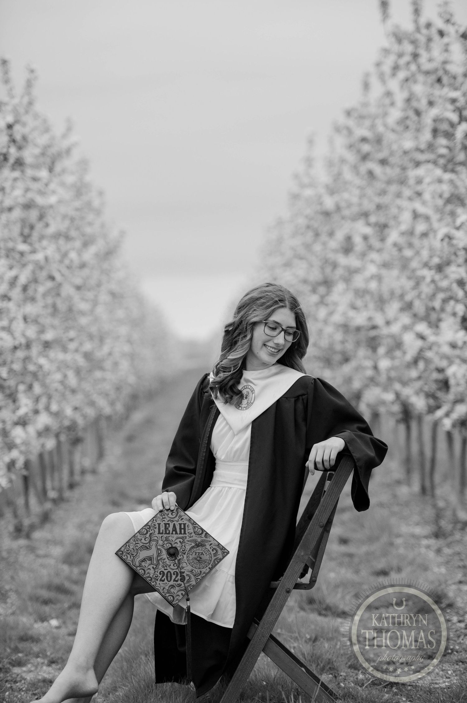 Graduate in navy cap and gown poses outdoors with blooming spring trees and a photographer's watermark visible.