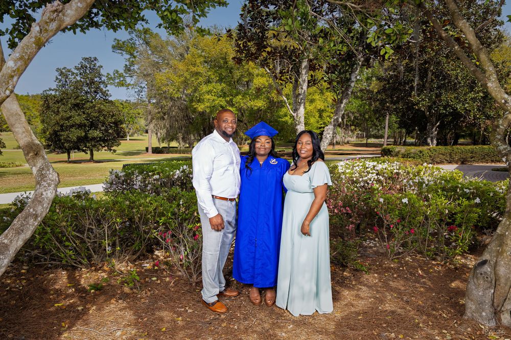Group photo in outdoor garden setting with person wearing blue graduation gown and white dress.