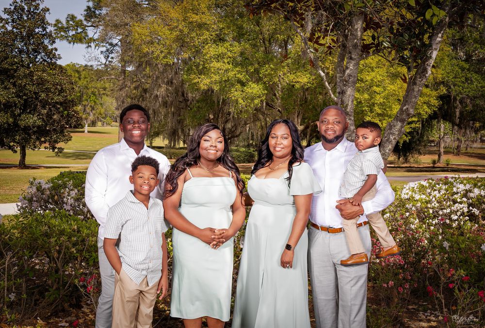Family in white and grey attire poses together in a garden setting with flowering bushes and trees.