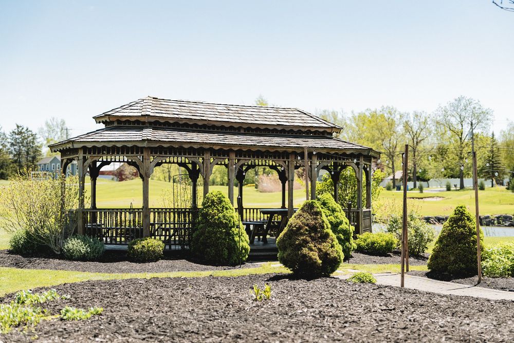 Wooden gazebo with shingled roof stands in landscaped garden area surrounded by trimmed bushes.
