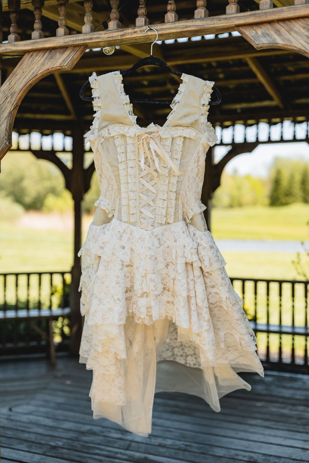 A vintage-style cream lace corset dress with ruffled layers hanging on display in a wooden gazebo.